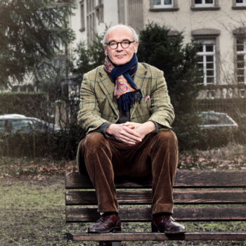Portrait von Martin Mosebach. er sitzt auf der Lehne einer Parkbank und blickt, die Hände auf den Knien verschränkt, in die Kamera.