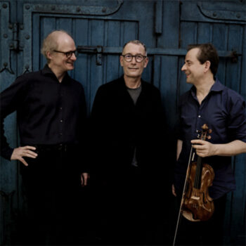 Drei Männer, die für ein Foto posieren. Der Mann in der Mitte blickt direkt in die Kamera, der Mann rechts und der Mann links lächeln sich an und sehen nicht direkt in ide Kamera. Der Mann rechts hält eine Violine sowie einen Bogen in der Hand.