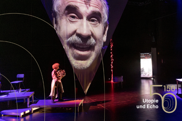 Das übergroße Portrait eines Mannes auf dunklen, nur von enigen Lichtpunkten erhellten Bühne.