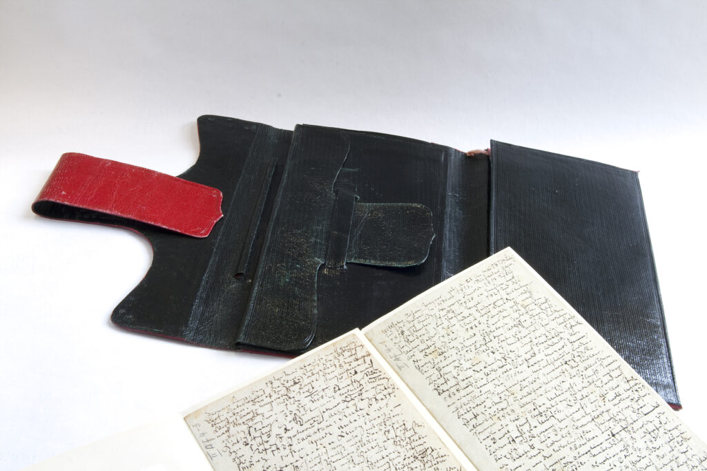 Das Bild zeigt eine schwarz-rote Brieftasche Richard Wagners auf weißem Untergrund, davor ein Notizbuch mit handschriftlichen Tagebucheinträgen, die der Komponist im Alter von 22 Jahren begann und die später zur Grundlage seiner Autobiographie wurden.